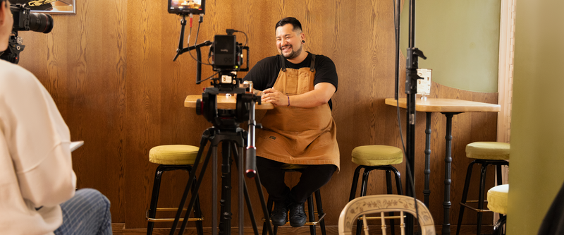 A man in a brown apron sits on a stool in a café, smiling while being filmed for an interview with cameras and lighting equipment set up around him.