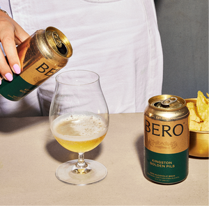 Person pouring BERO Kingston Golden Pils beer into a glass with a can and chips on a table.