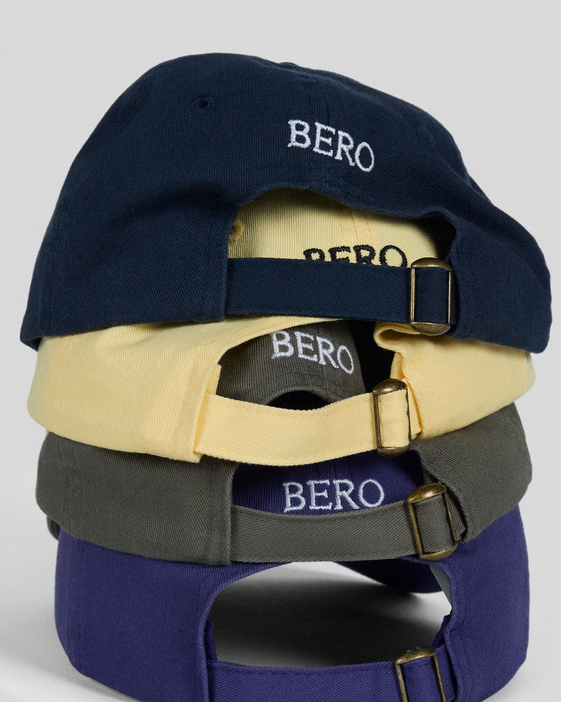 Stack of four BERO baseball caps in navy, cream, olive, and purple with adjustable straps