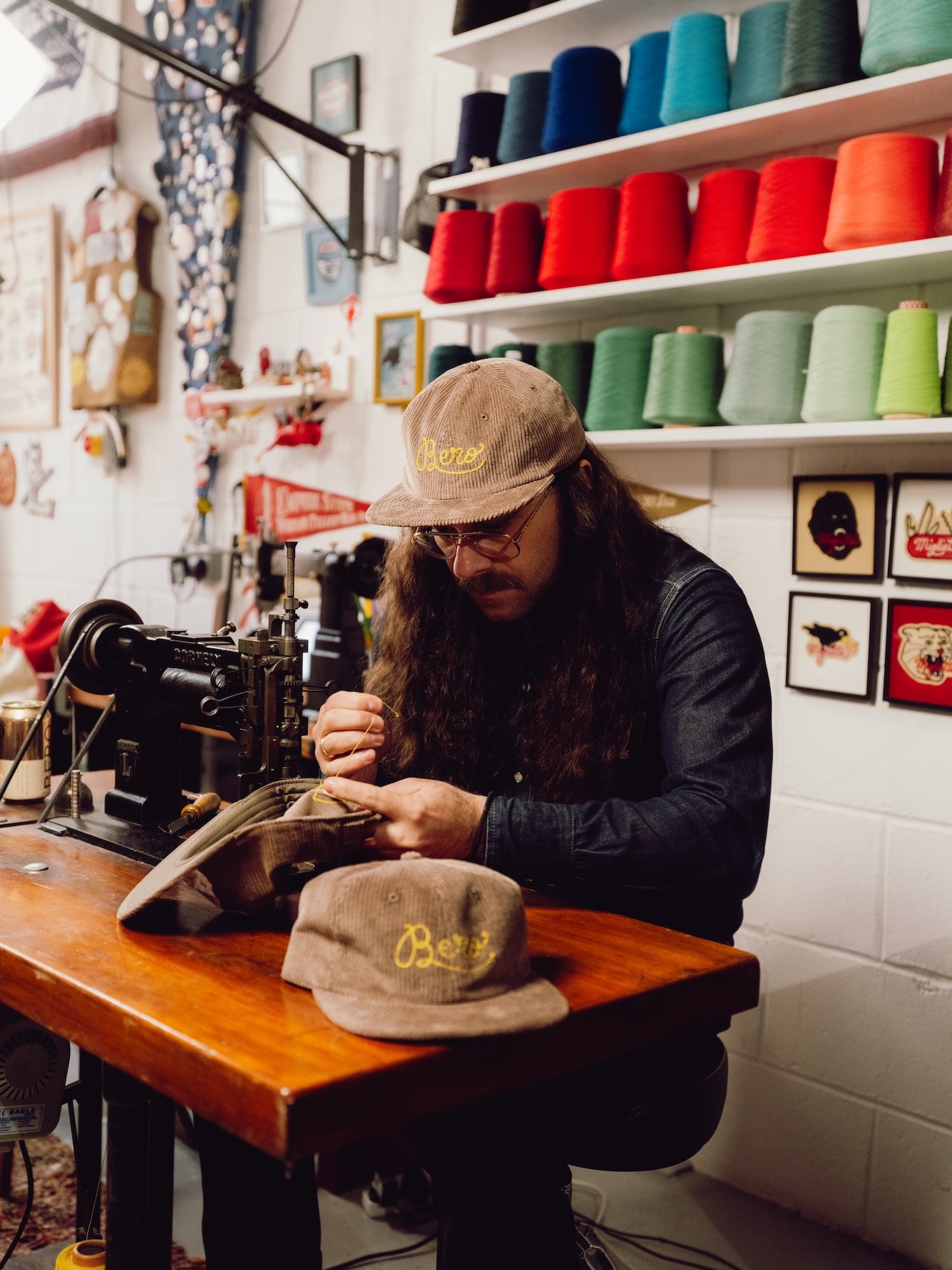 Giulio Miglietta working on a sewing machine in a workshop with colorful spools of thread on shelves.