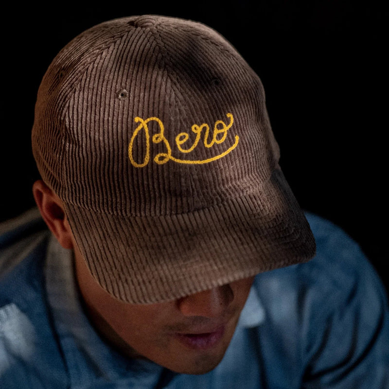 Person wearing brown corduroy baseball cap with yellow BERO logo embroidered on front, looking down in blue shirt
