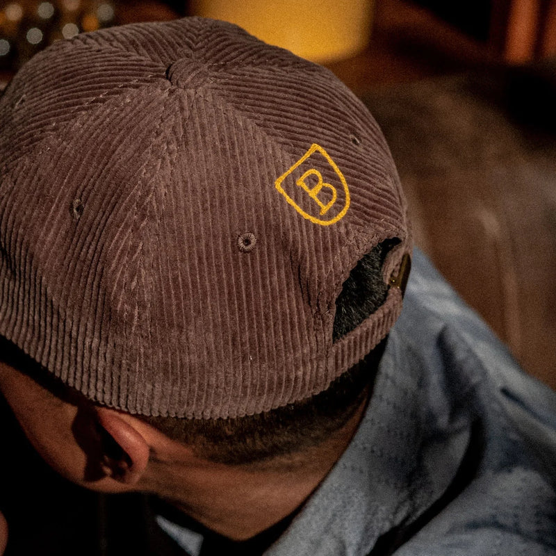 Brown corduroy baseball cap with yellow embroidered BERO logo worn by person in gray hoodie