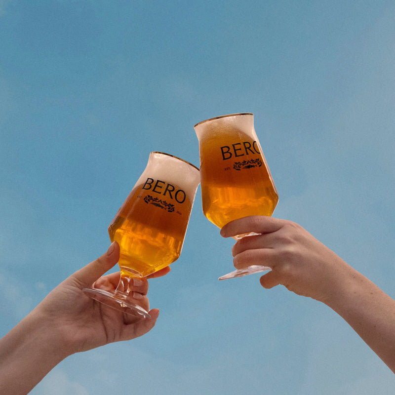 Two hands toasting with BERO beer glasses filled with golden non-alcoholic beer against blue sky