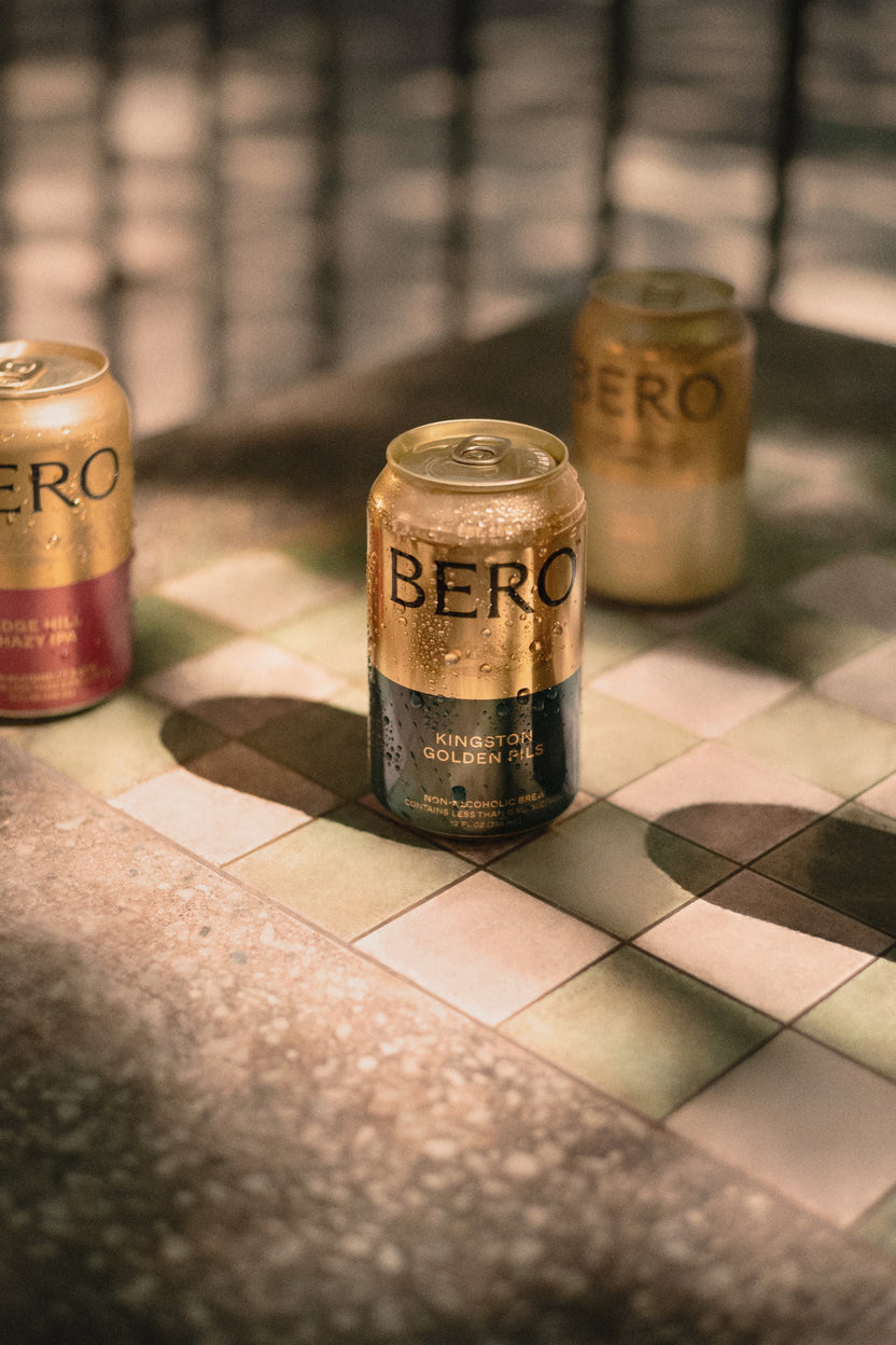 Three cans of Bero beer on a checkered tile surface with a blurred background