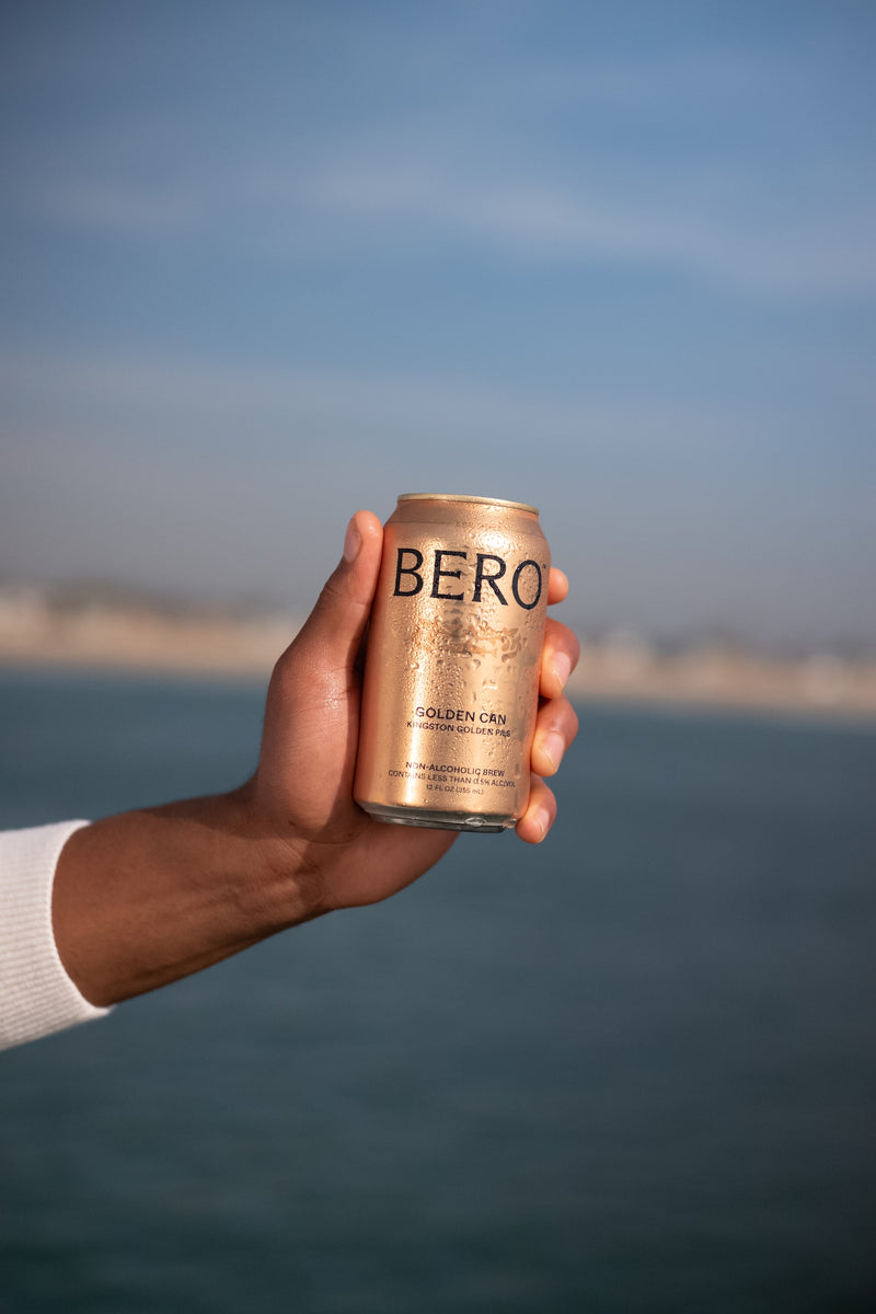 Hand holding a gold BERO non-alcoholic beer can against a blurred ocean and sky background on a sunny day