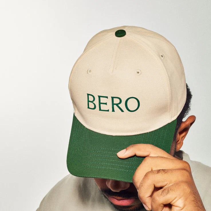 Person wearing cream and green BERO baseball cap, hand raised to brim, against white background