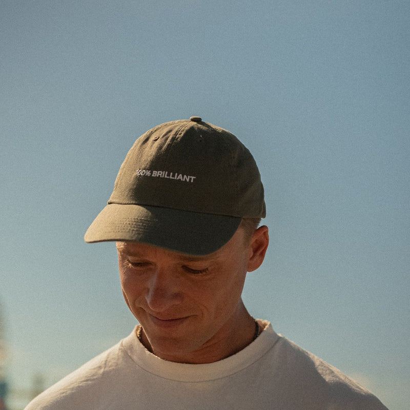 Person wearing olive green baseball cap with "100% BRILLIANT" text, looking down with gentle smile