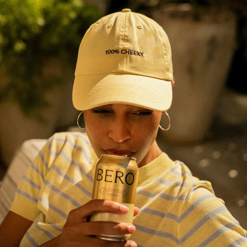 Person wearing "100% CHEEKY" baseball cap drinking from BERO non-alcoholic beer can