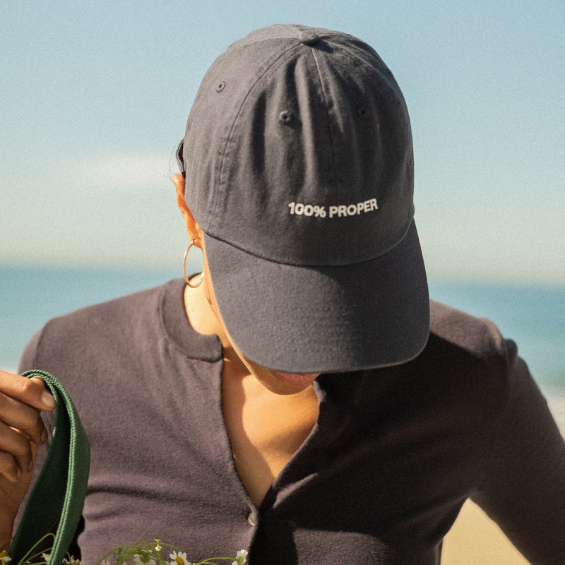 Person wearing dark "100% PROPER" baseball cap outdoors, holding green rope or cord