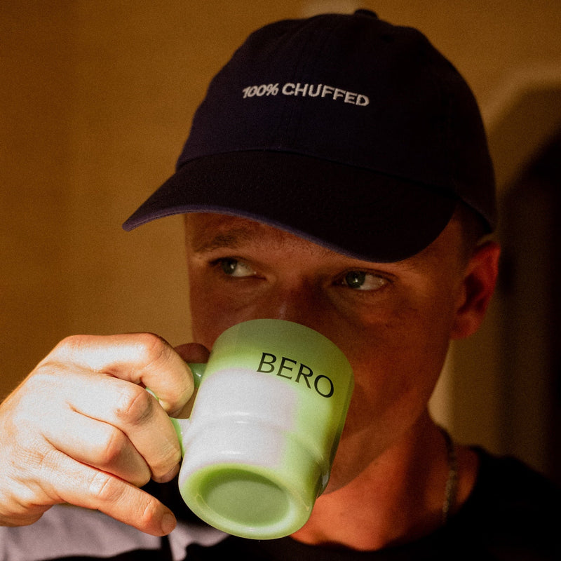 Person wearing black "100% CHUFFED" cap drinking from green BERO branded mug