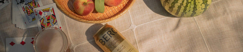 BERO non-alcoholic beer can on checkered blanket with playing cards, fruit basket, and watermelon