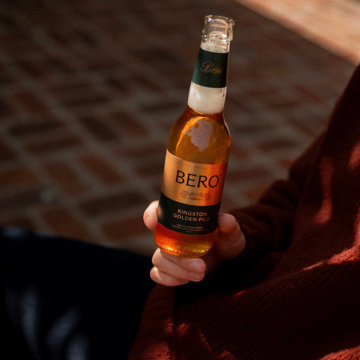 A hand holding a bottle of Bero, showing the amber liquid and label, with a person wearing a rust-colored knit sweater in warm sunlight against a brick background.