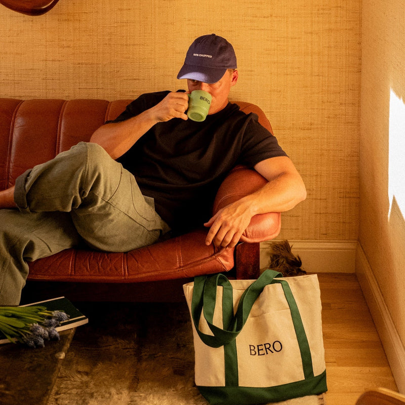 Man in baseball cap drinking from green BERO mug while relaxing on leather sofa, with BERO tote bag on floor beside him
