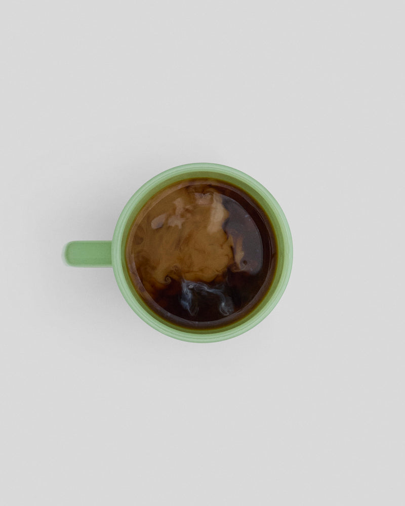 Overhead view of a light green ceramic mug filled with dark brown beer, showing foam and bubbles on the surface