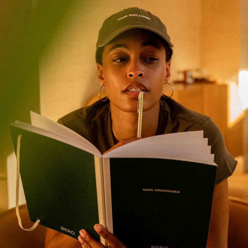 Person reading open BERO "100% Organized" notebook with pen in mouth, wearing cap and hoop earrings