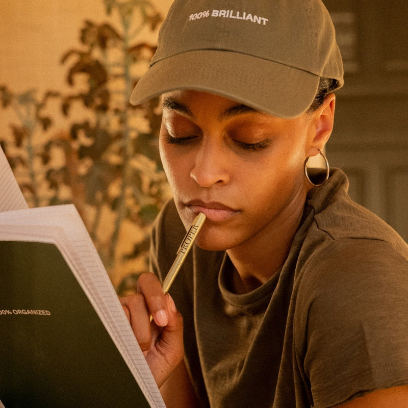 Person in olive cap and hoodie thoughtfully holding pen while reading from notebook