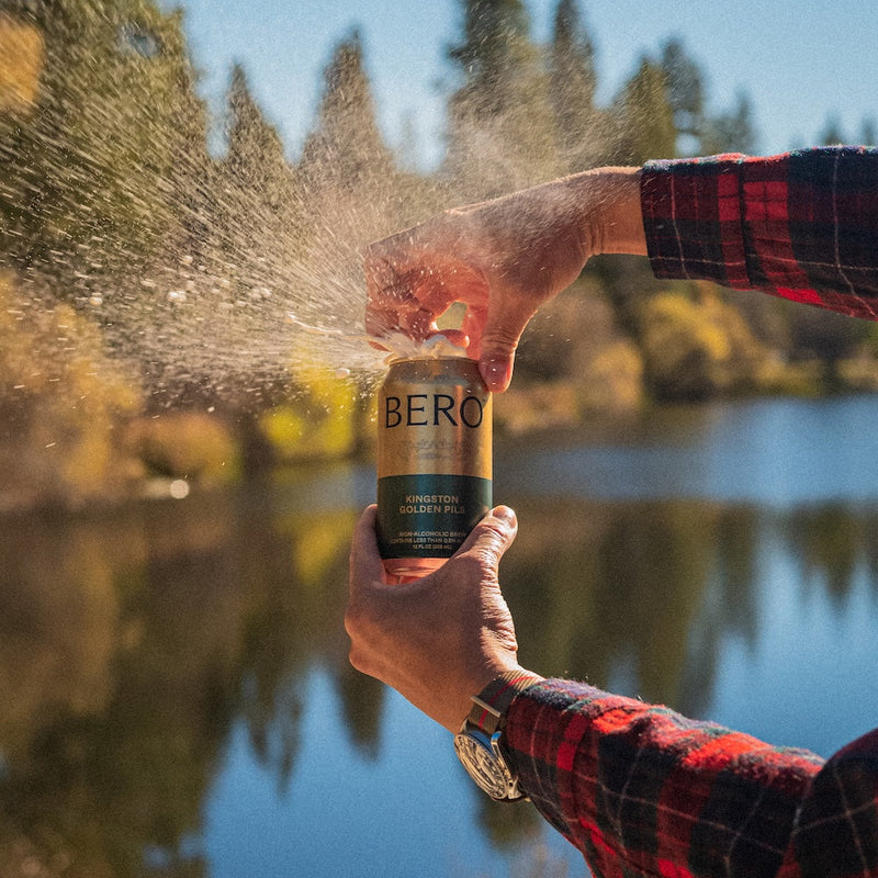 Person in red flannel shirt holding BERO Kingston Golden Pils can outdoors by a lake with water splashing in sunlight